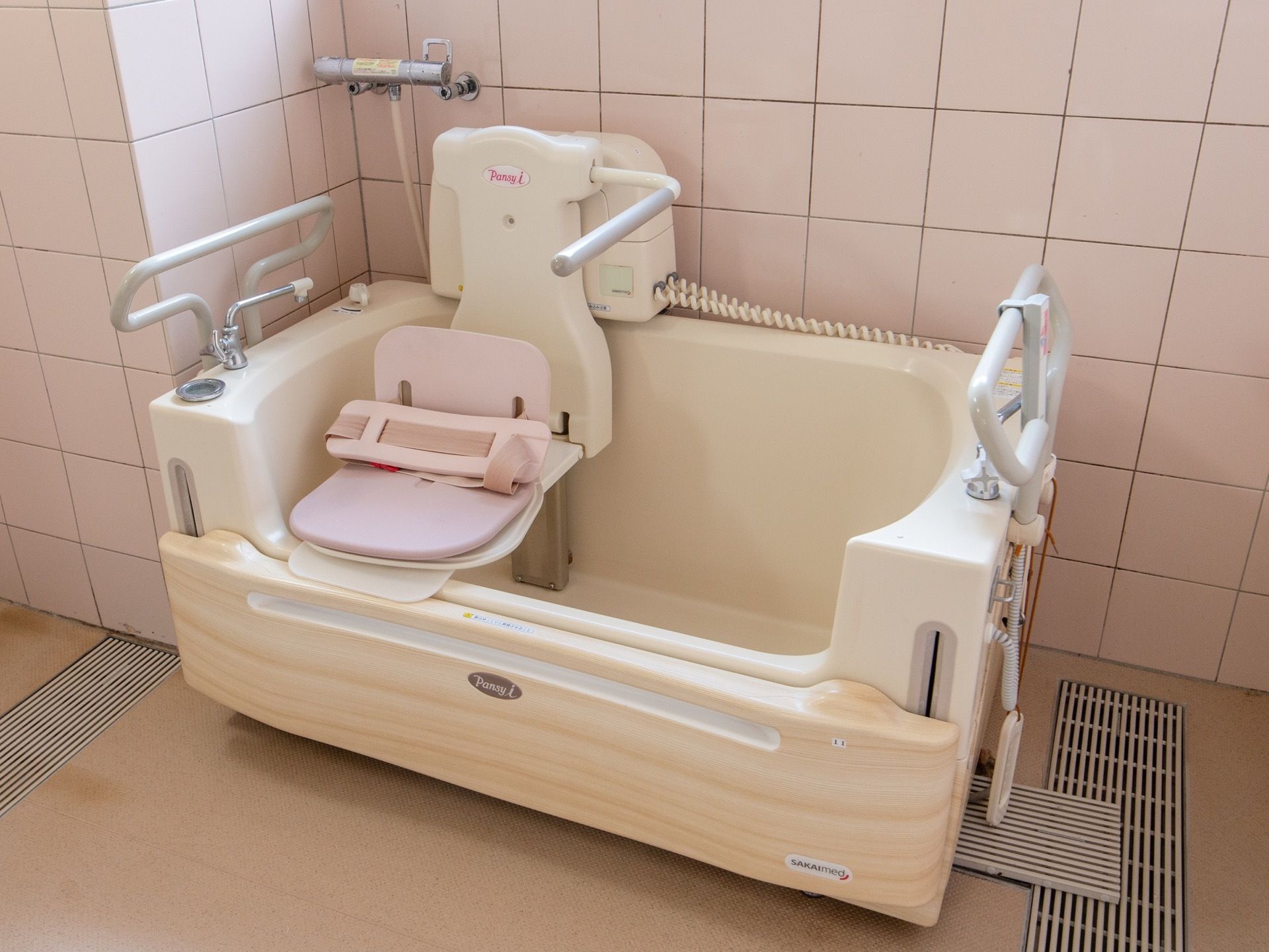 ピンクのタイルに囲まれた浴室がある。内部に可動式の椅子が取りつけられた、入浴介護用の浴槽が置かれている。