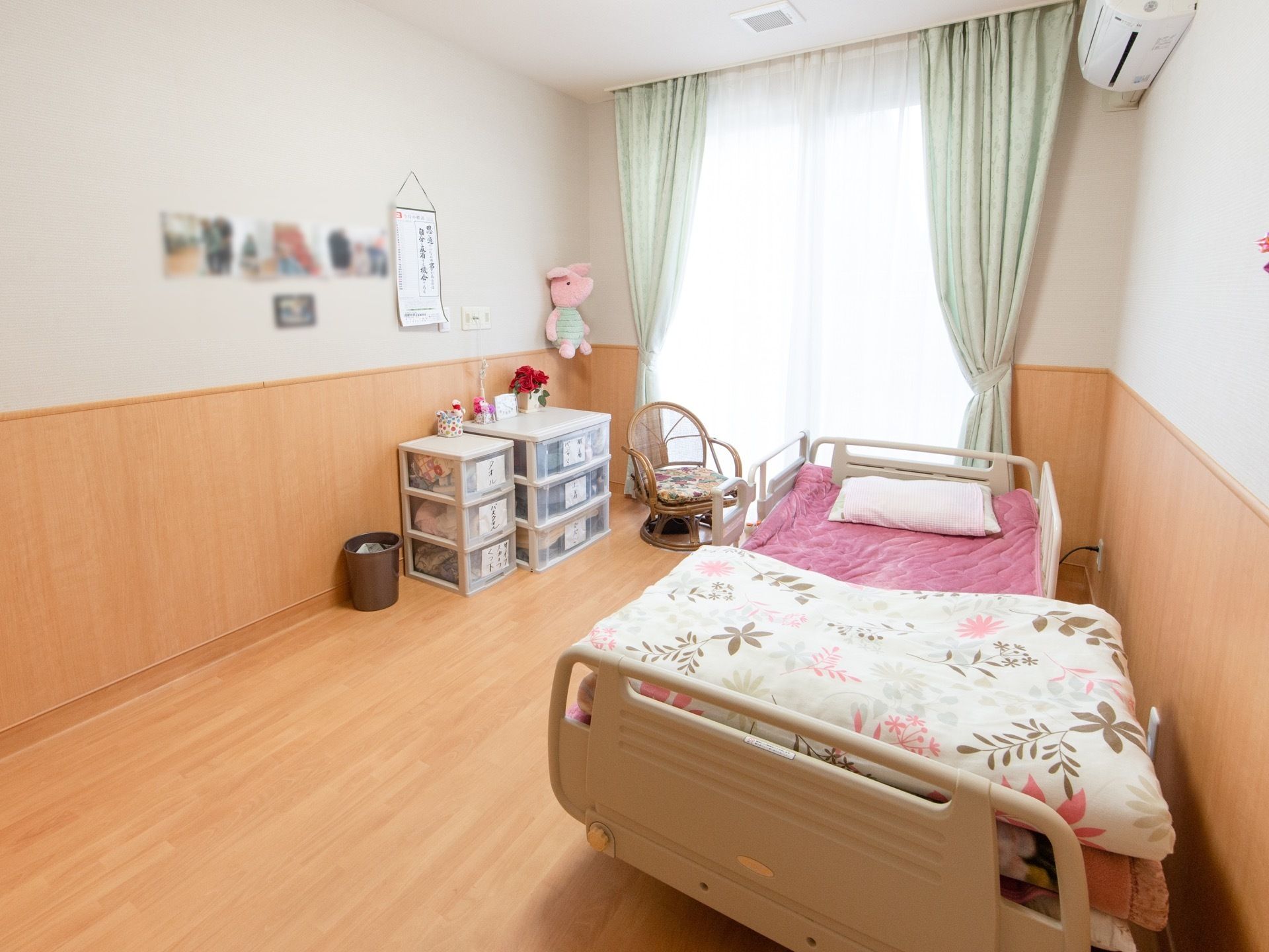 大きな窓からの日当たりが良い、明るい居室がある。ライトブラウンのフローリングで、壁の半分も同じ素材を使っている。介護用ベッドが置かれている。