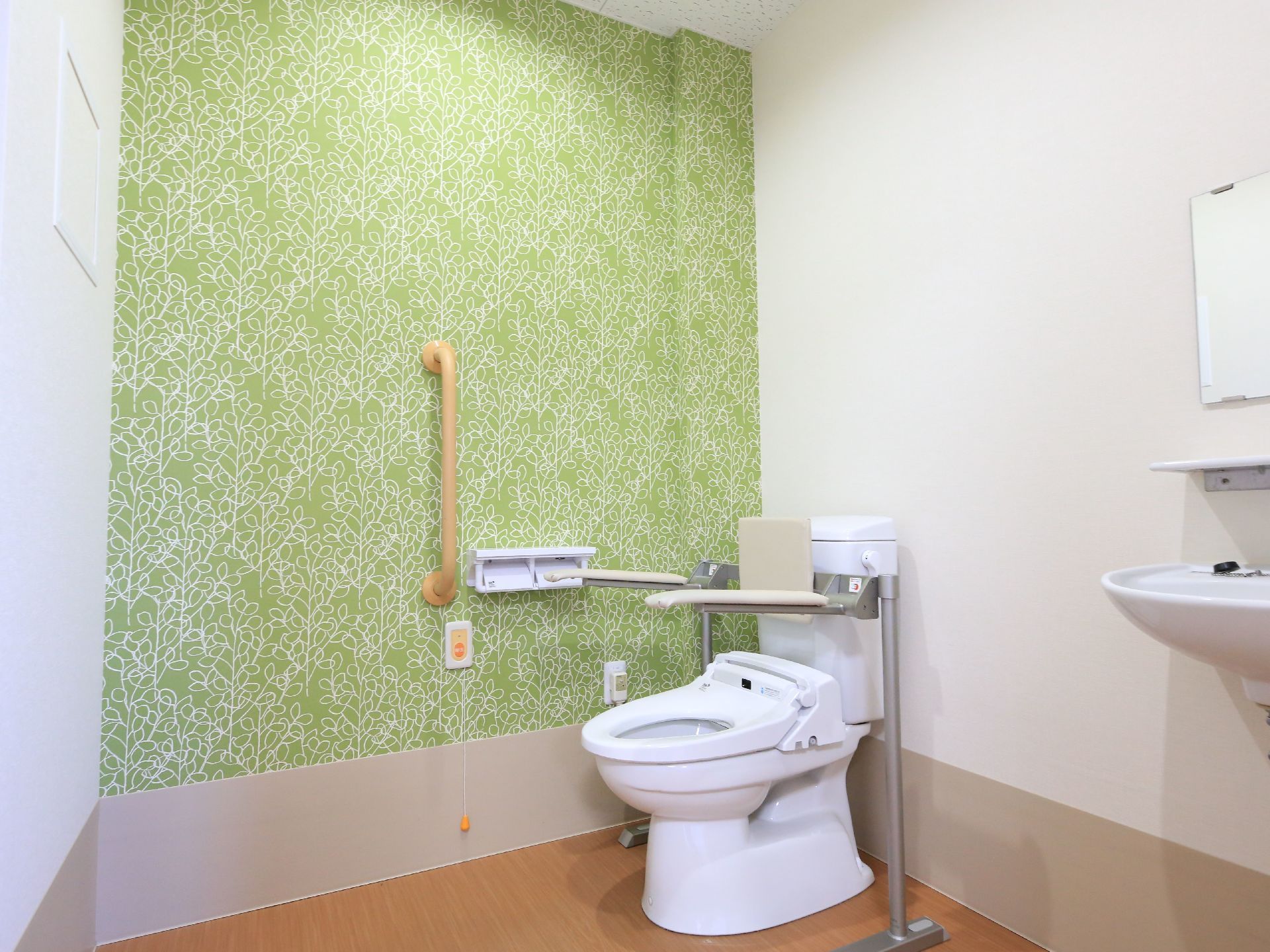 トイレには背もたれ、肘掛けがついている。便座は蓋がないので、アクションが少なく立ち座りできる。緊急ボタンも横に設置してある。