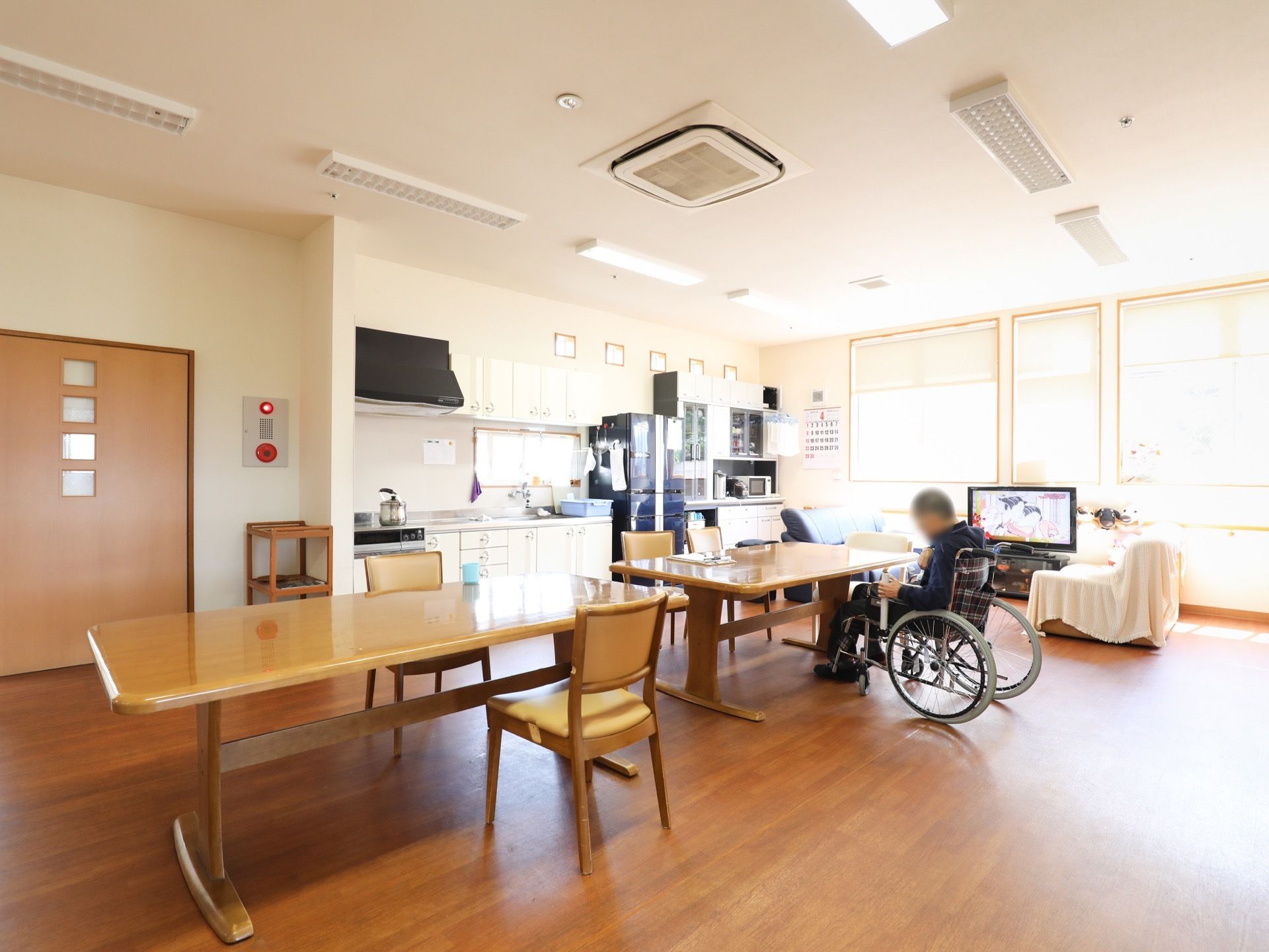 部屋の中央に食卓が2台並んでいる。壁には台所が設けられている。窓の付近にはテレビが置かれ、その近くにソファが配置されている。