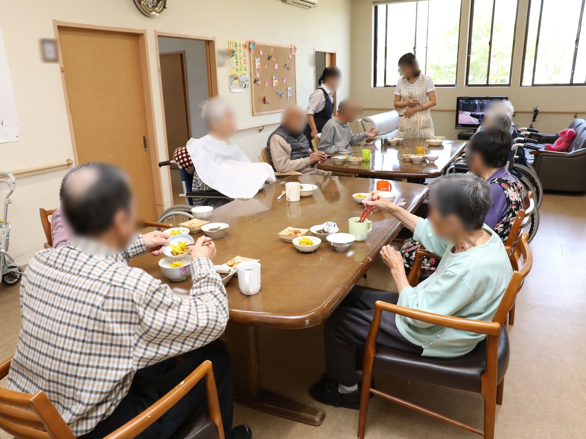 室内の中心に食卓が2台、両端を合わせて置かれている。窓の近くにテレビ台が設置されており、茶色系の一人用ソファも配置されている。