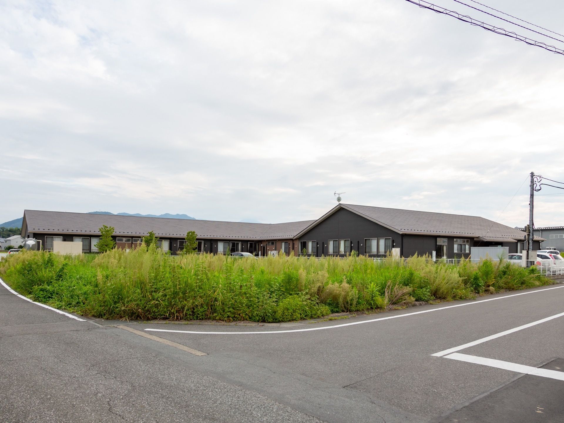 アスファルトの道路の奥に雑草が生えた敷地があり、その奥に平屋建ての黒い建物がある。建物の奥に山が見え、空は曇っている。