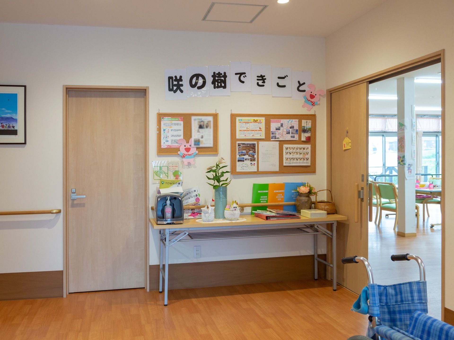 木目のフローリングの床の上に折り畳みの車椅子がある。壁際に花瓶などの雑貨が置かれた長テーブルが置いてある。