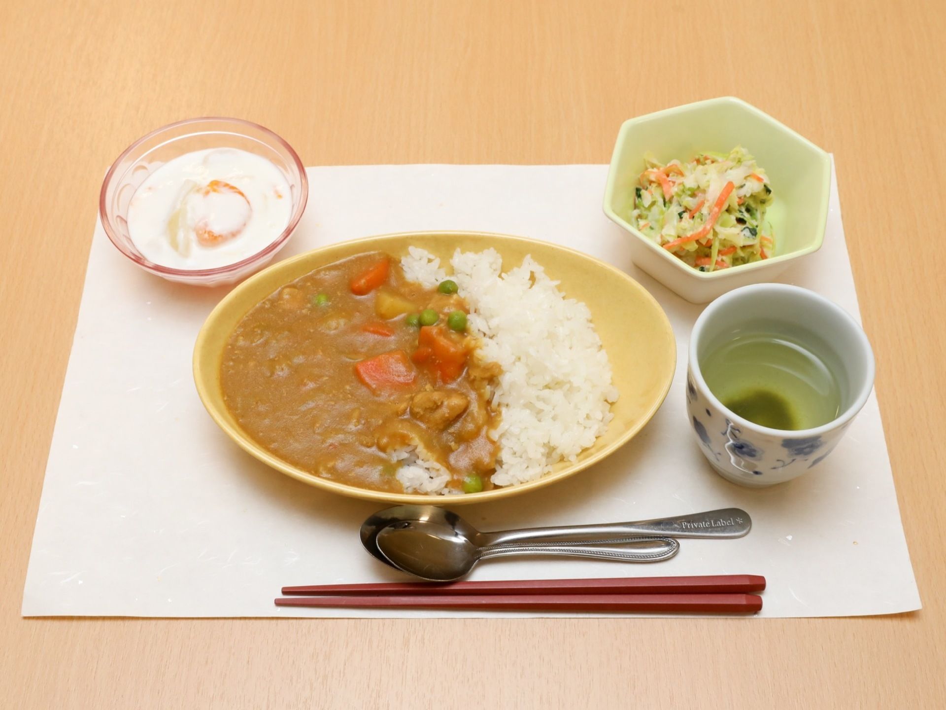 白い紙の上に、食事が並べられている。メインはカレーライスで、サラダとデザートも添えられている。お茶も置かれている。