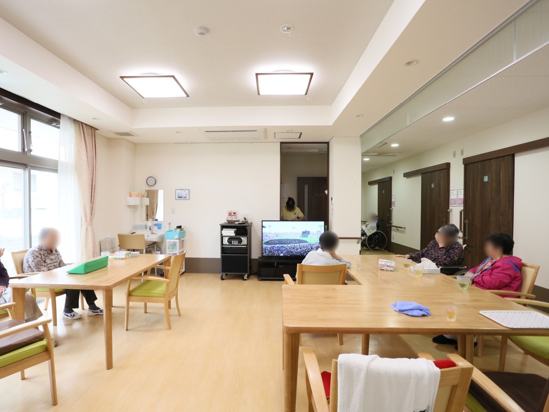 明るい色の木製テーブルを複数配置している。入居者様が椅子に座りお茶を飲んだり、テレビを見たりしてくつろいでいる。