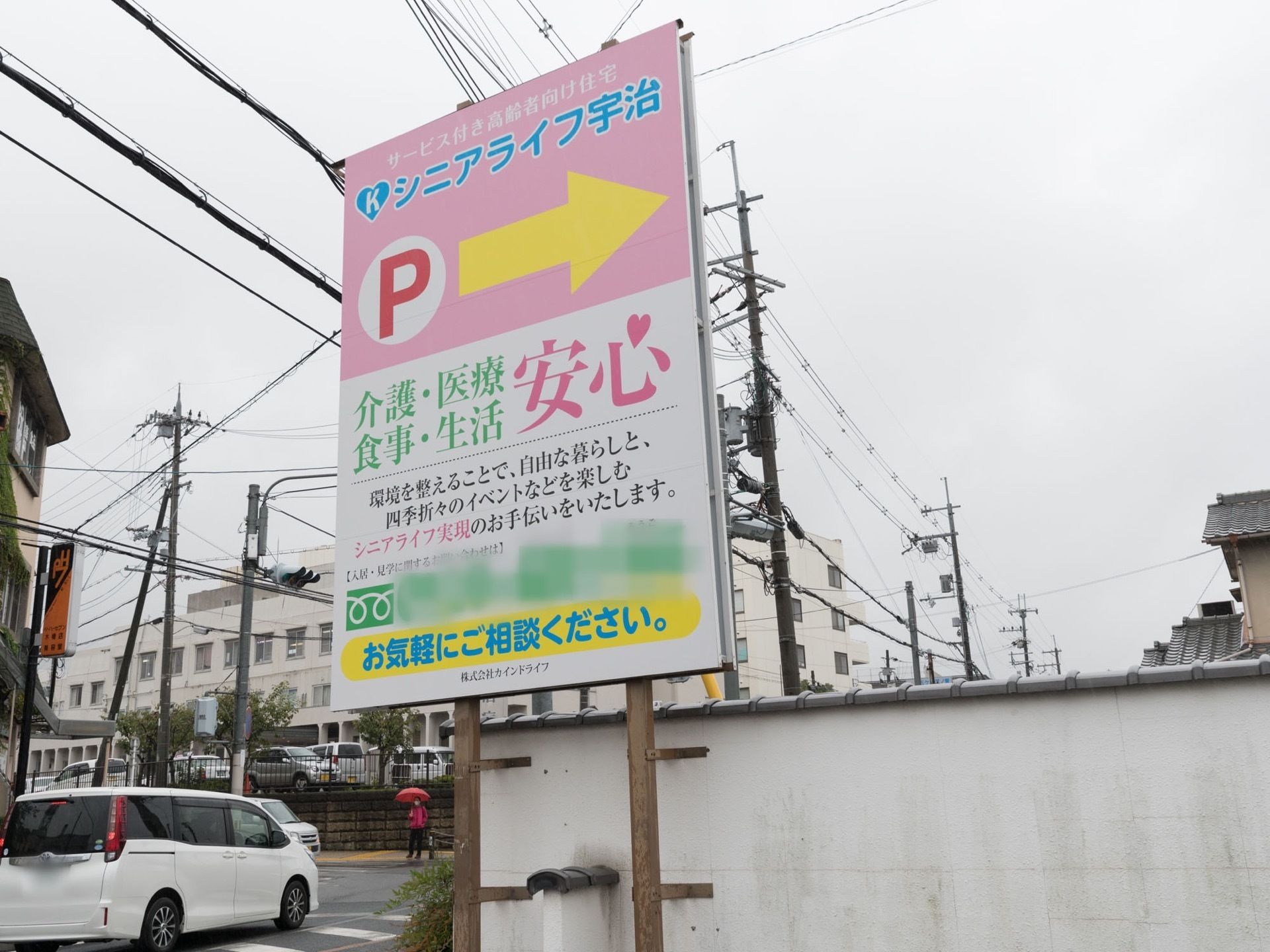 道路沿いにある白い外壁の前には施設の看板がある。看板には施設名やパーキングの場所、電話番号などが書かれている。