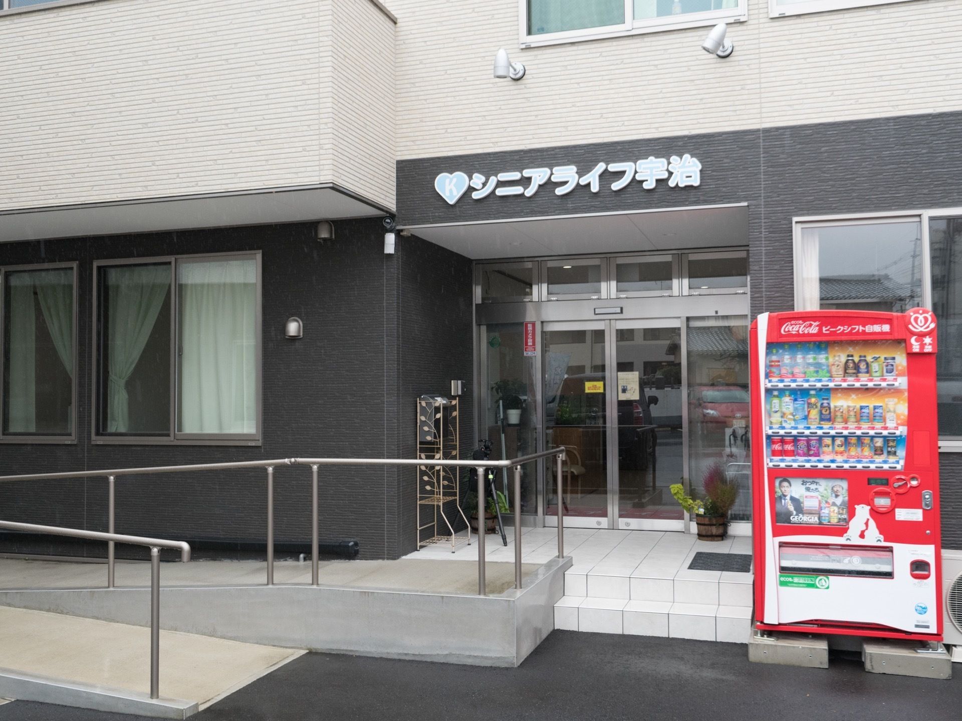自動販売機が置かれている。施設入口には階段と緩やかなスロープがある。施設名が入り口の上部に書かれている。