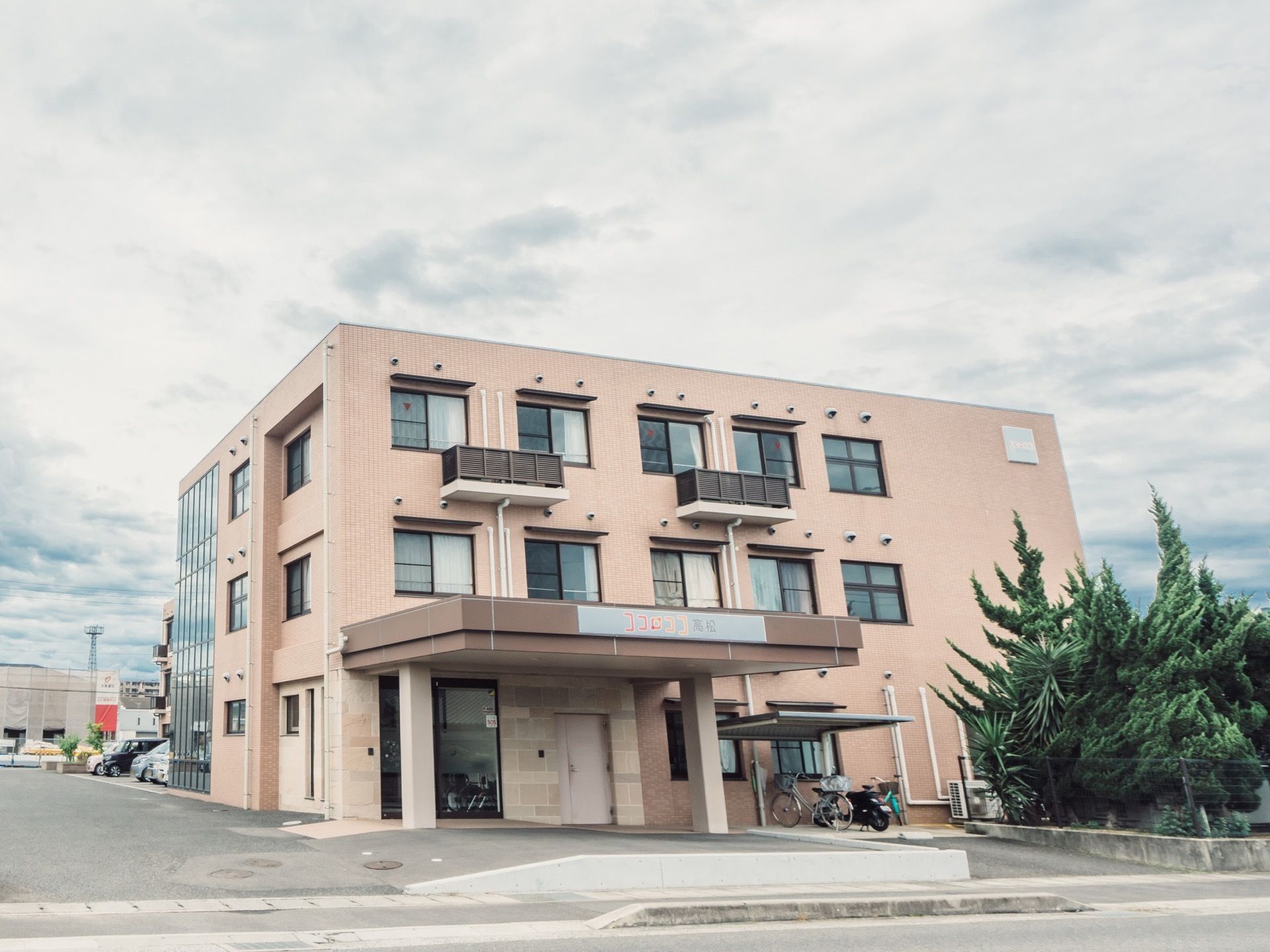 柔和な雰囲気の３階建ての施設。建物の周囲はアスファルトで舗装されており、玄関の脇に常緑樹が茂っている。