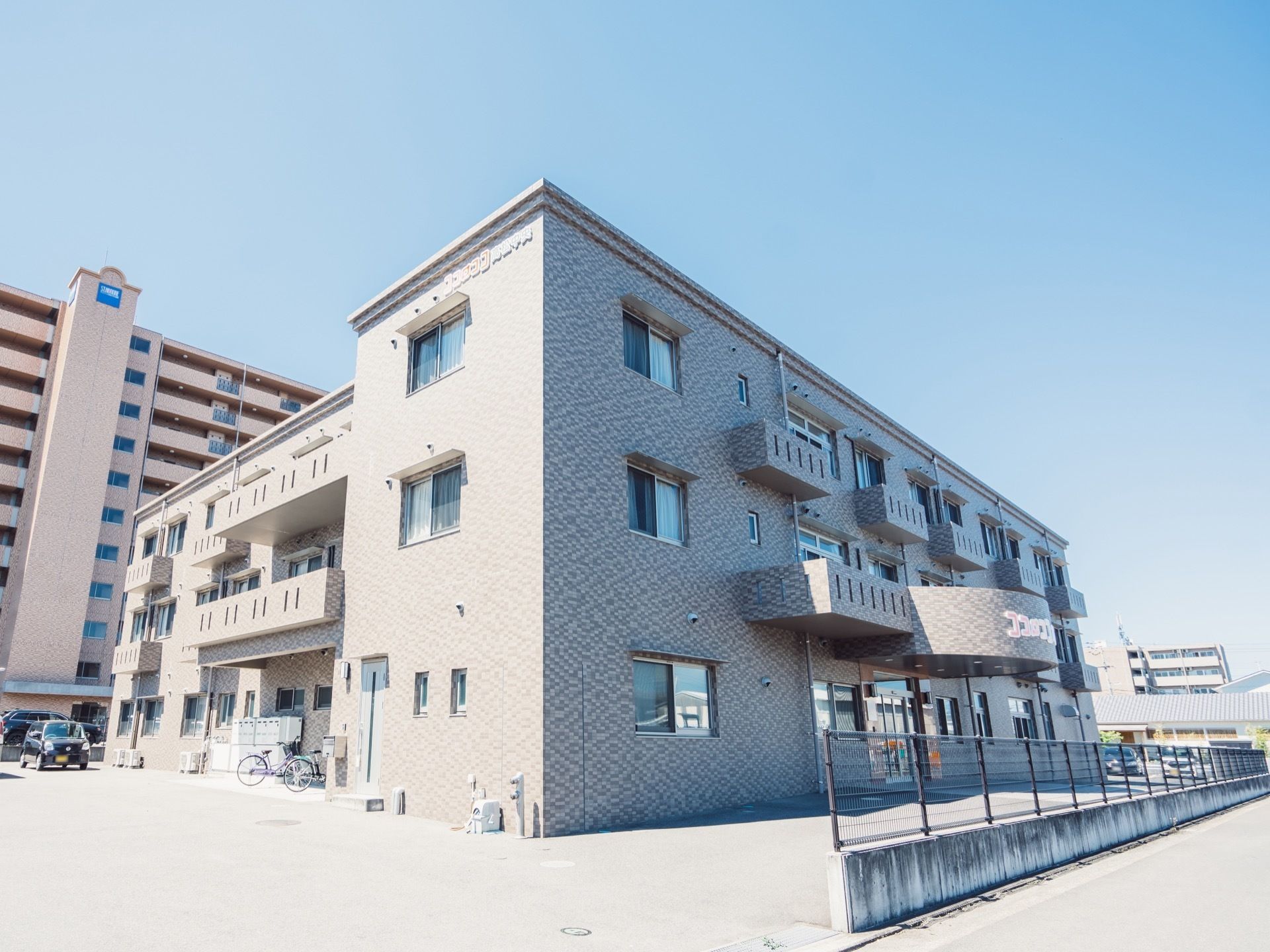 施設はおしゃれな外観で3階建てとなっている。敷地は広々しており、道路側にはフェンスが建てられている。