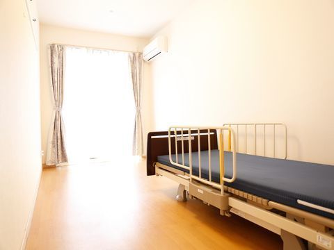洋室タイプの居室には介護用のベッドが1つ置かれている。奥には窓がありカーテンも備え付け。壁にはエアコンが設定されている。