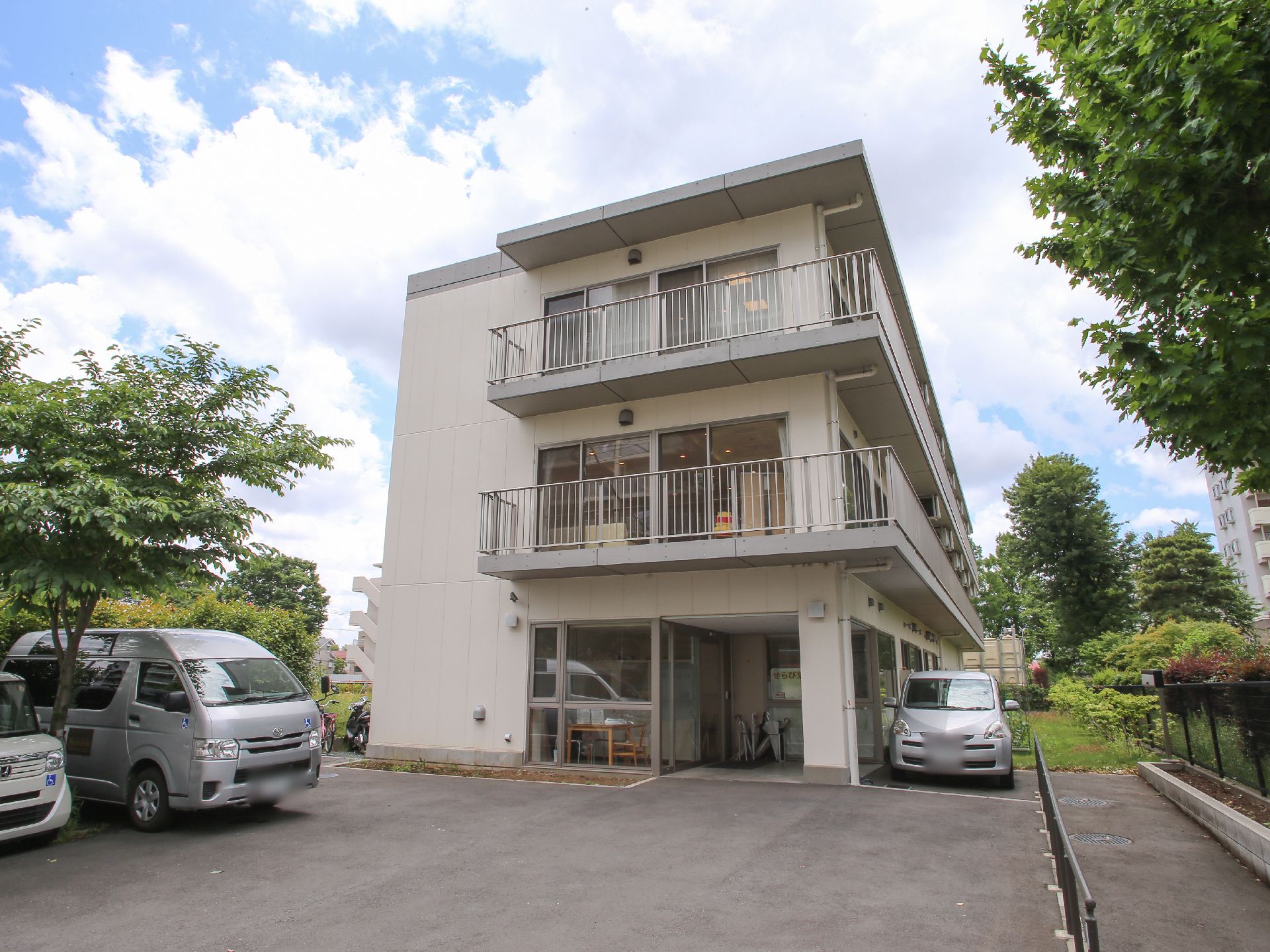 施設は白壁の三階建でアパートのような外観になっている。手前には駐車場が設けられていて車が数台停まっている。周囲には木々があり緑豊か。