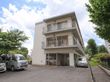 施設は白壁の三階建でアパートのような外観になっている。手前には駐車場が設けられていて車が数台停まっている。周囲には木々があり緑豊か。