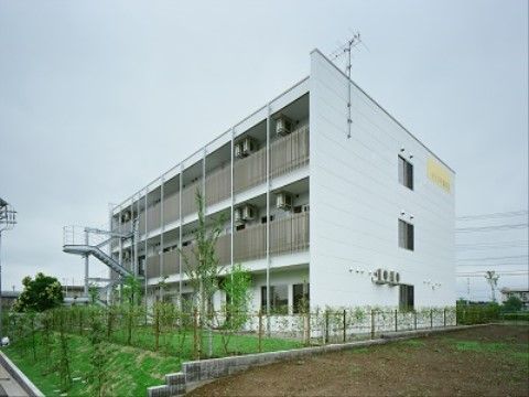 ３階建ての施設はシンプルな長方形のデザインの白い建物である。バルコニーに面した敷地は植物の緑で覆われている。