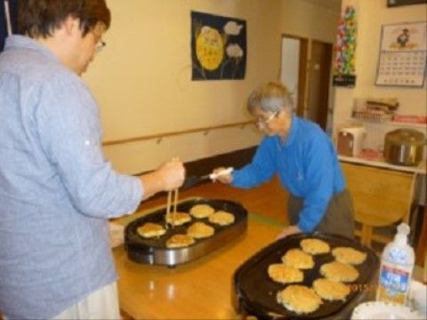 ホットプレートでお好み焼きを焼くスタッフと、それを手伝う入居者様の模様