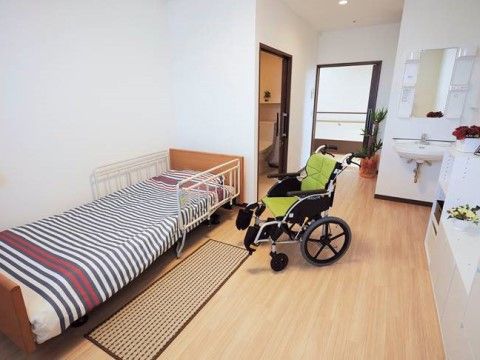 白を基調とした居室である。ベッドには手すりが付いている。部屋のところどころに小さな鉢が置かれており、綺麗な花が咲いている。