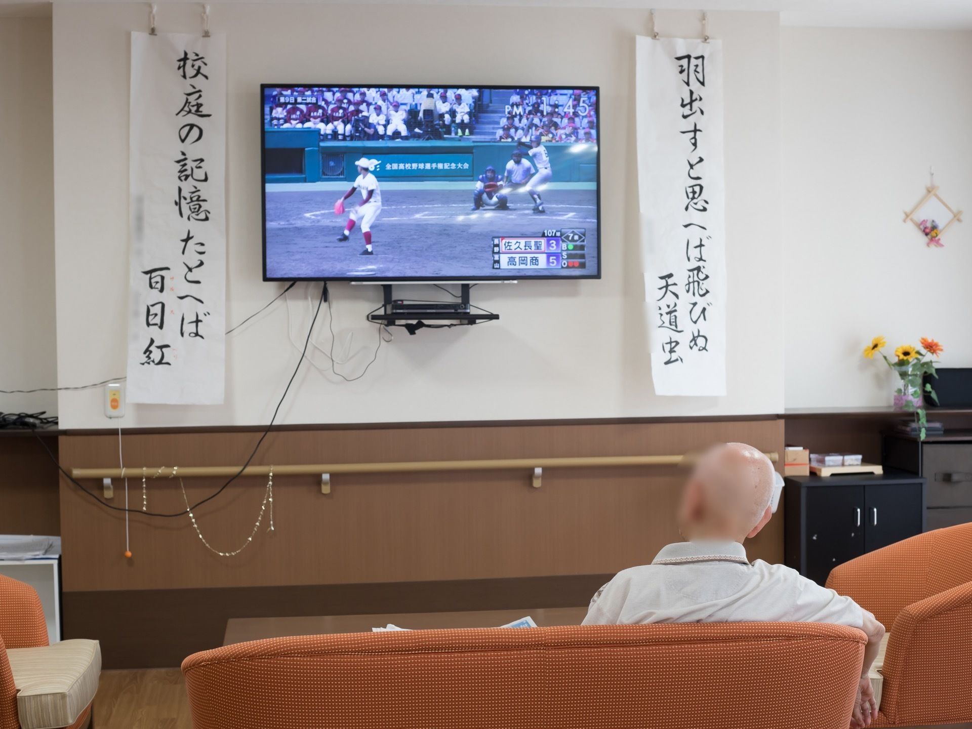 大型のテレビが壁の上方に設置されている。男性がオレンジ色のソファでくつろぎながら高校野球を観ている。