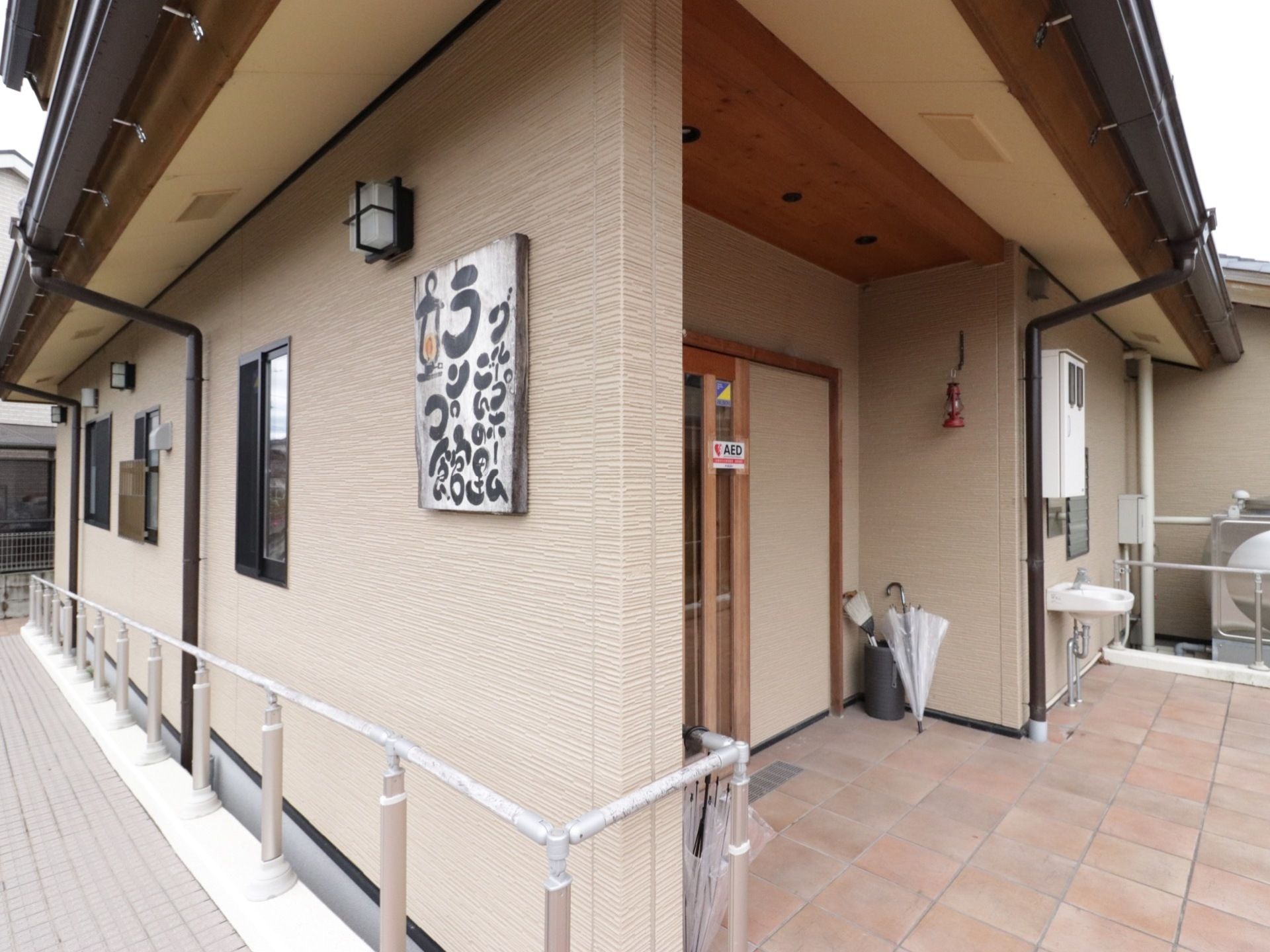 玄関横の壁に、毛筆の手書き風の施設名が書かれた看板が付いている。その壁に沿って、手すりとスロープが設置されている。