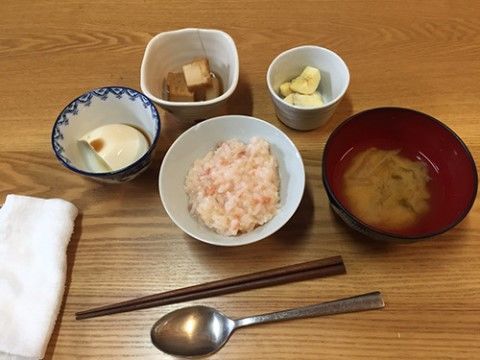 木目のテーブルの上に料理が並べられている。ご飯とみそ汁、小鉢に入った豆腐やバナナなどがある。箸とスプーン、お手拭きが置いてある。