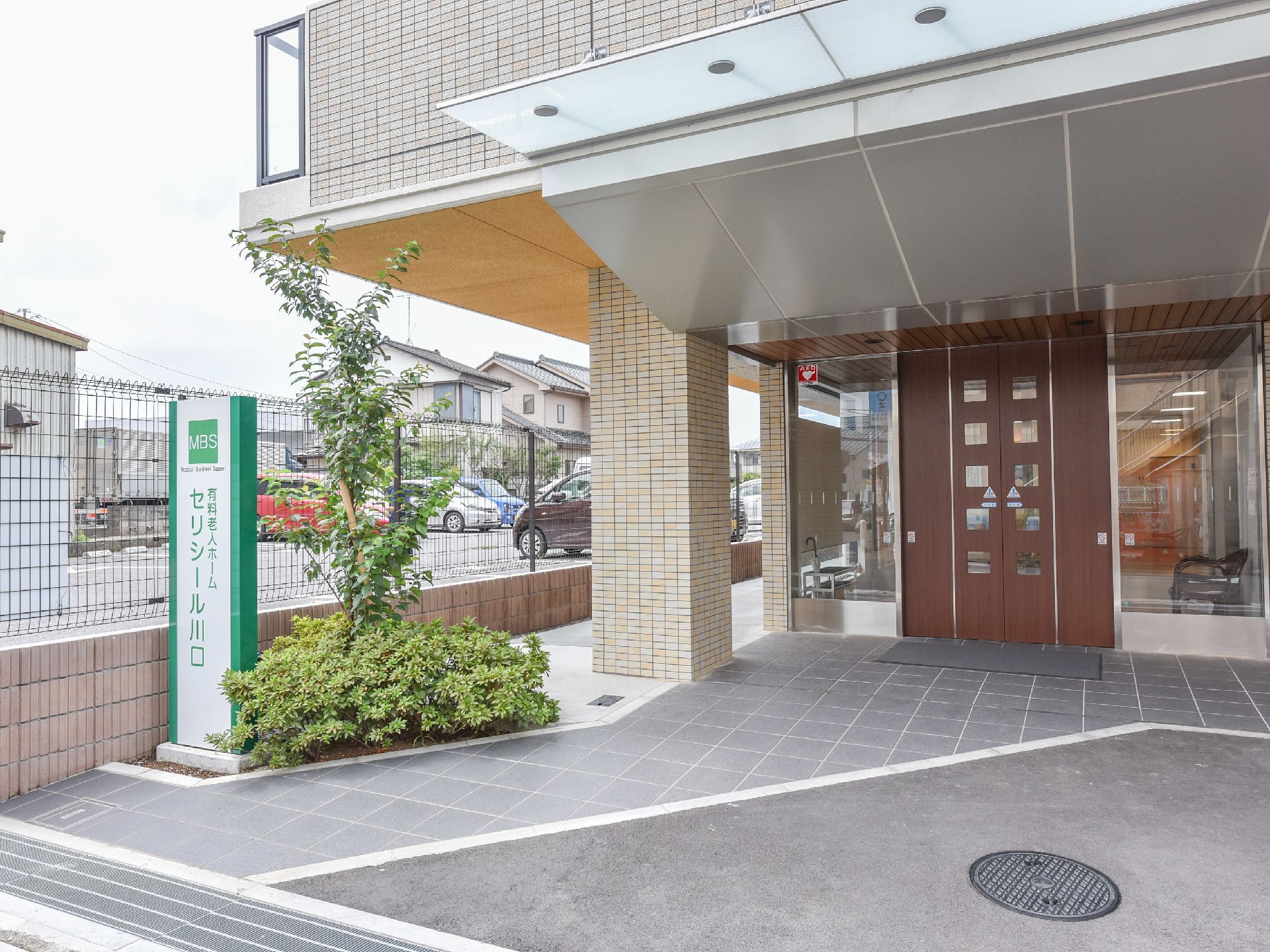 施設の入り口には左側に植え込みがあり、その手前に施設名が書かれた看板が立っている。足元はタイル敷きで玄関前まで続いている。