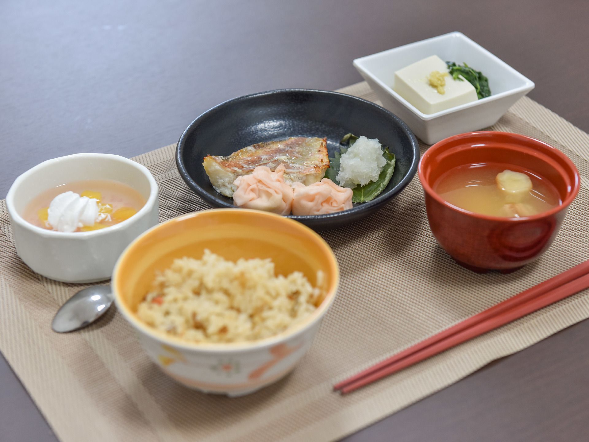 ベージュのランチョンマットの上に1人分の食事がセットされている。ご飯と味噌汁のほか、おかずが2皿とデザートがある。