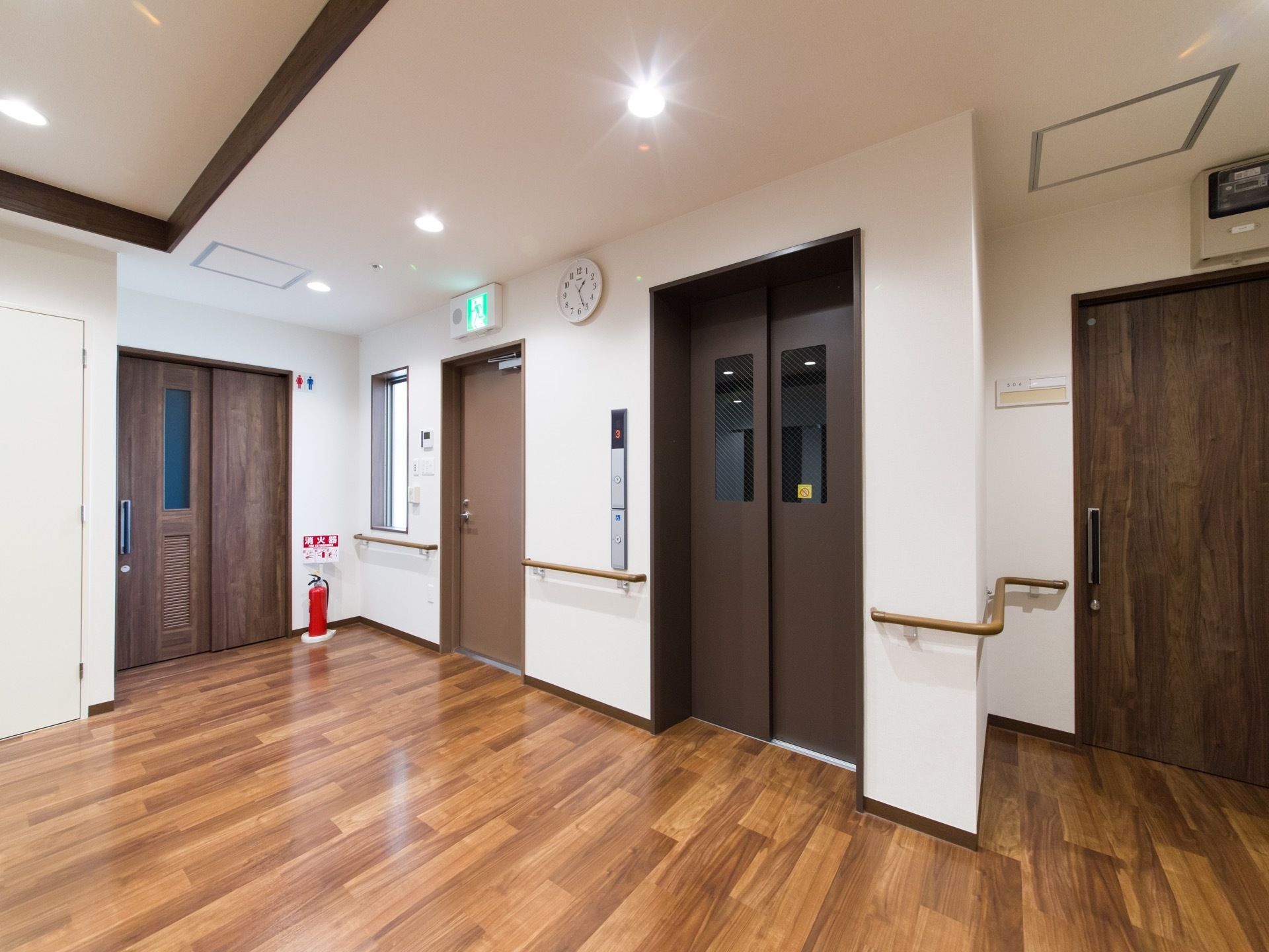 エレベーターの隣には非常階段、トイレがある。消火器も用意されている。壁には手すり、置時計が設置されている。