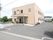 二階建てのベージュの建物がある。施設のすぐ横が駐車場になっている。敷地内は舗装されており、一部に砂利が敷かれている。
