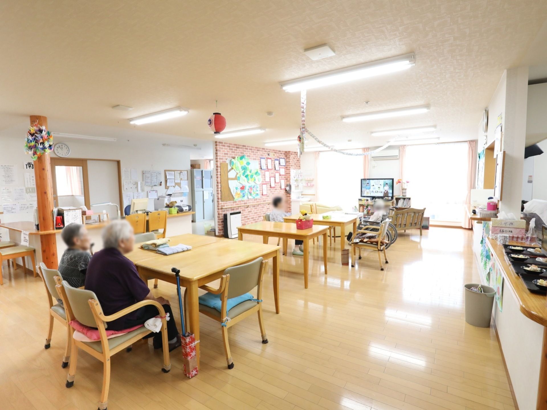明るい食堂には四角いテーブルと椅子がいくつか並んでいる。カウンターには食事が並び、壁には入居者様の作品が飾られている。