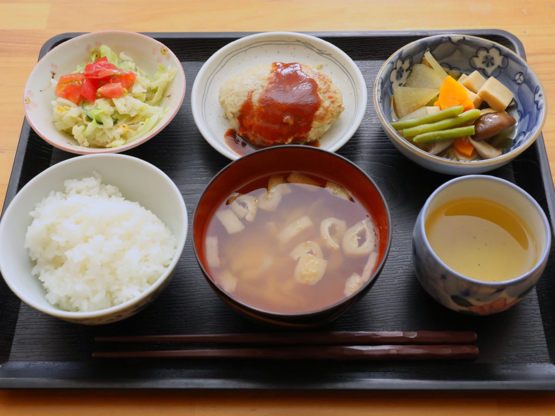 黒塗りのお盆に乗った美味しそうな食事。小鉢に一品ずつ盛られ、サラダ、ハンバーグ、煮物、ご飯とお味噌汁が並べられている。緑茶の湯飲みもある。