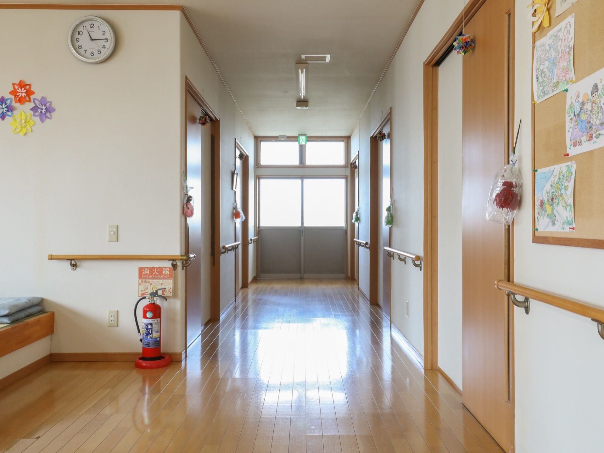 廊下の両サイドに木製の手すりが取りつけられている。突き当たりには非常口があり、壁に設置されたボードには絵が貼られている。