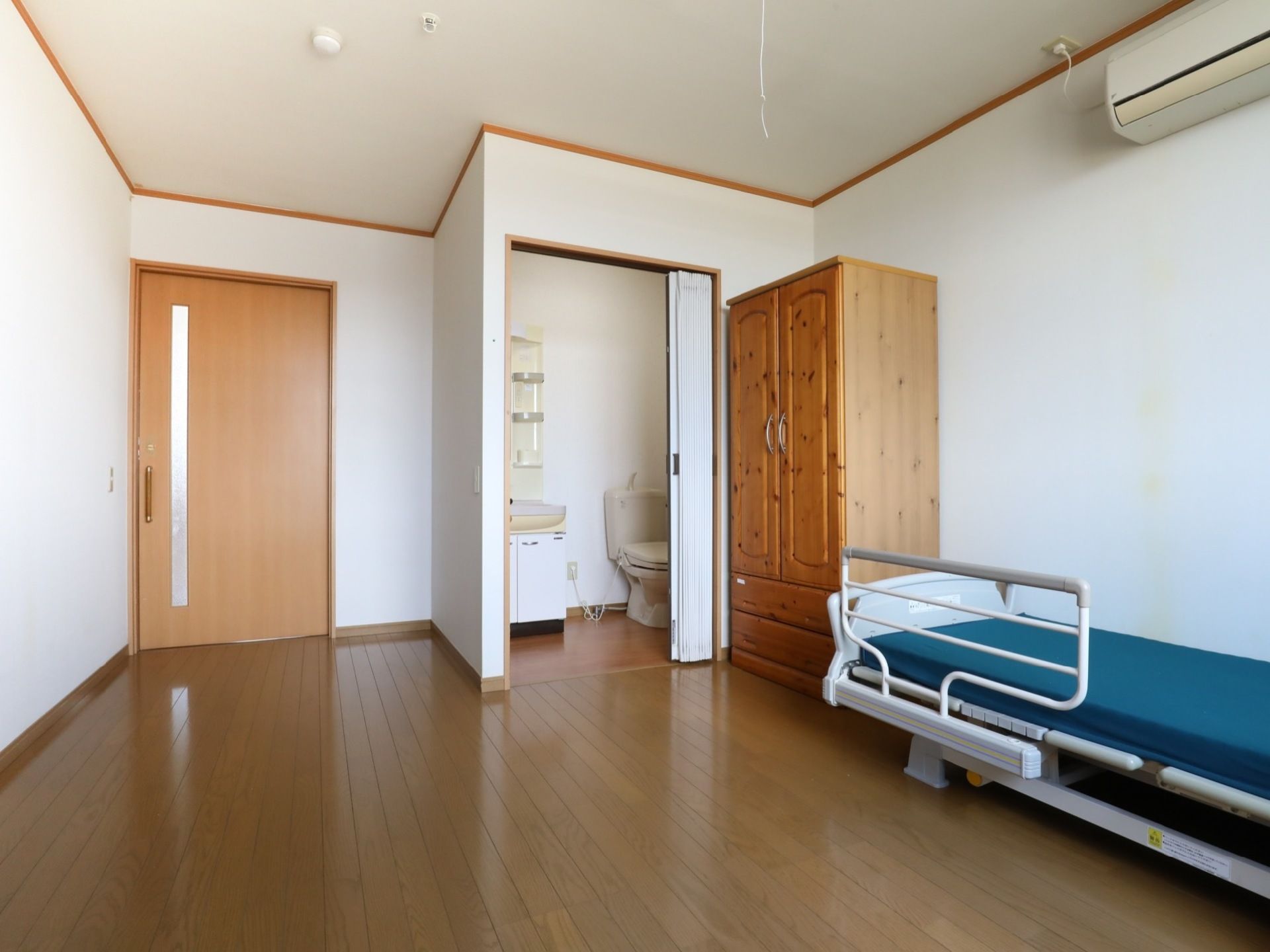 トイレや洗面台、エアコンが備えつけられた居室。フローリングの床の上には、木製の収納棚や介護ベッドが置かれている。