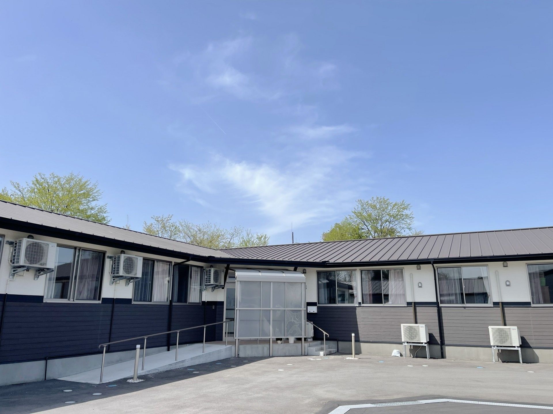 ベージュの淡い色彩の外観である。周辺は、閑静な緑豊かな住宅街で、施設の建物前には駐車スペースを備えている。