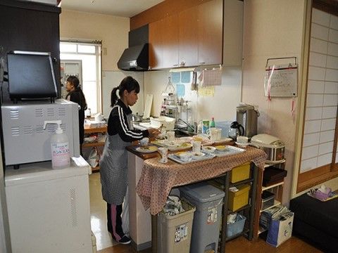 正面にキッチンカウンターがあり、食事トレーが数枚並んでいる。エプソンを付けた女性スタッフが盛り付けをしている。