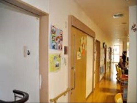 木製の床の廊下が続いている。左側の壁に横開きのドアが並んでいる。右側に入居者様が座っている様子が見える。