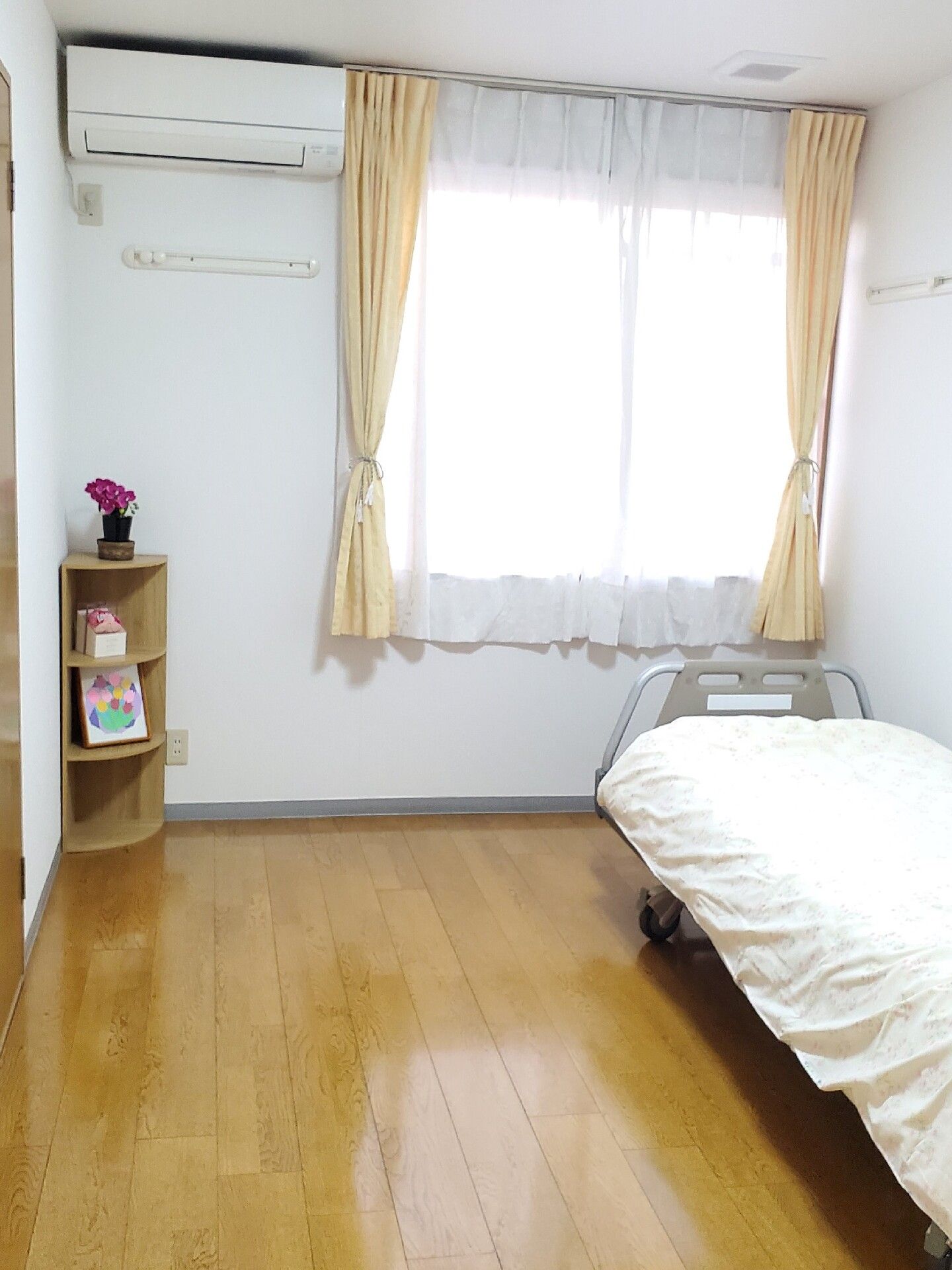 木製のフローリングの部屋にベッドなどが置かれている。左側には椅子の上にテレビが置かれている。大きな窓があって明るい。