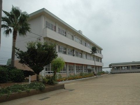 ３階建ての校舎で広いグラウンドがある。奥には体育館も見える。花壇には白い花が咲いており、シュロの木も植えられている。