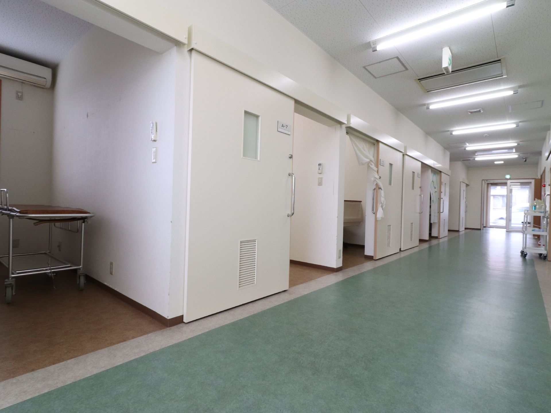緑色をした床の廊下がある。廊下には蛍光灯が取り付けられている。廊下の両サイドには部屋があり、一部には暖簾がかかっている。