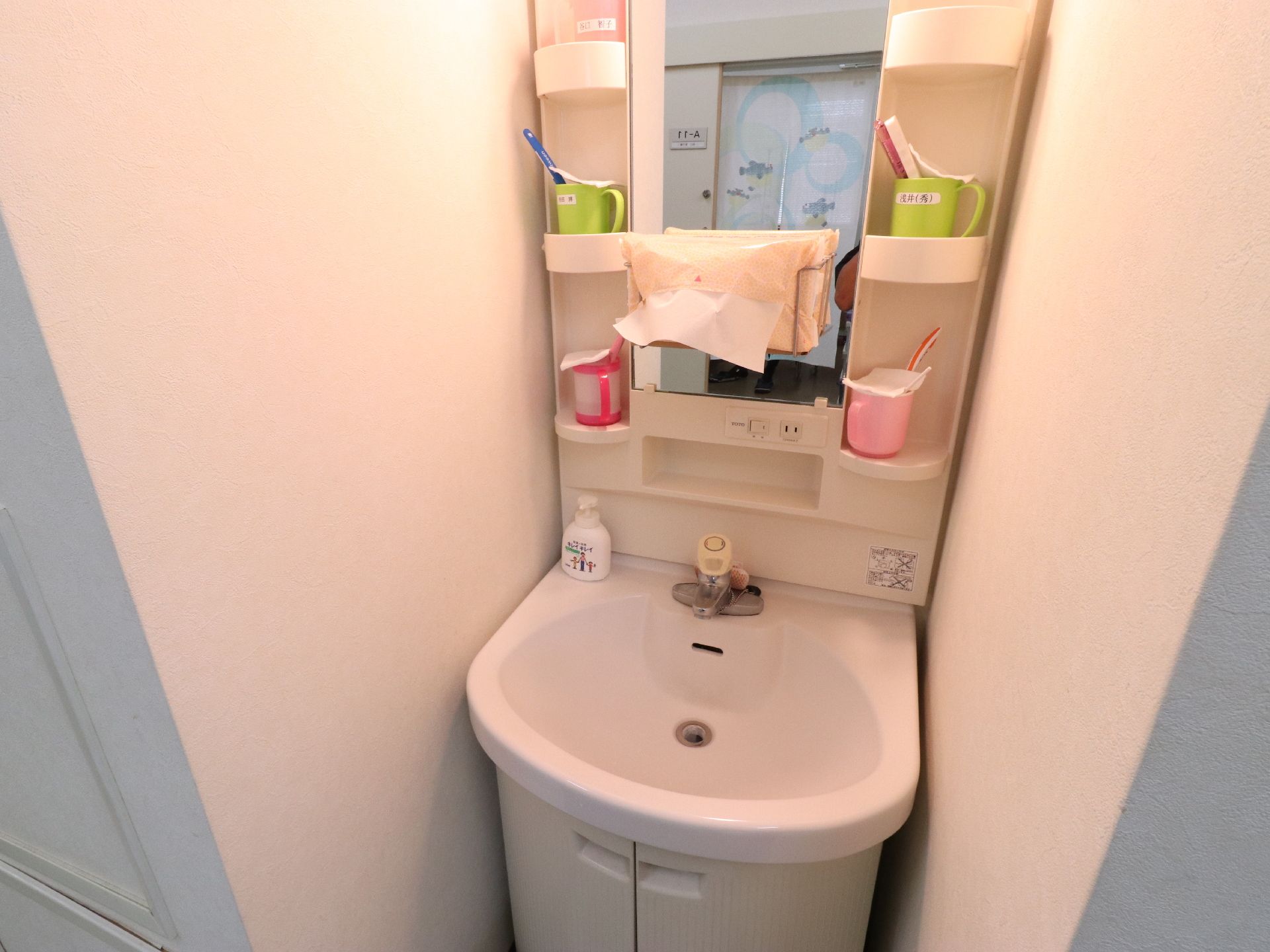 洗面台にはハンドソープが置かれている。収納部分には黄緑色と薄いピンク色、濃いピンク色のコップと歯ブラシが置かれている。