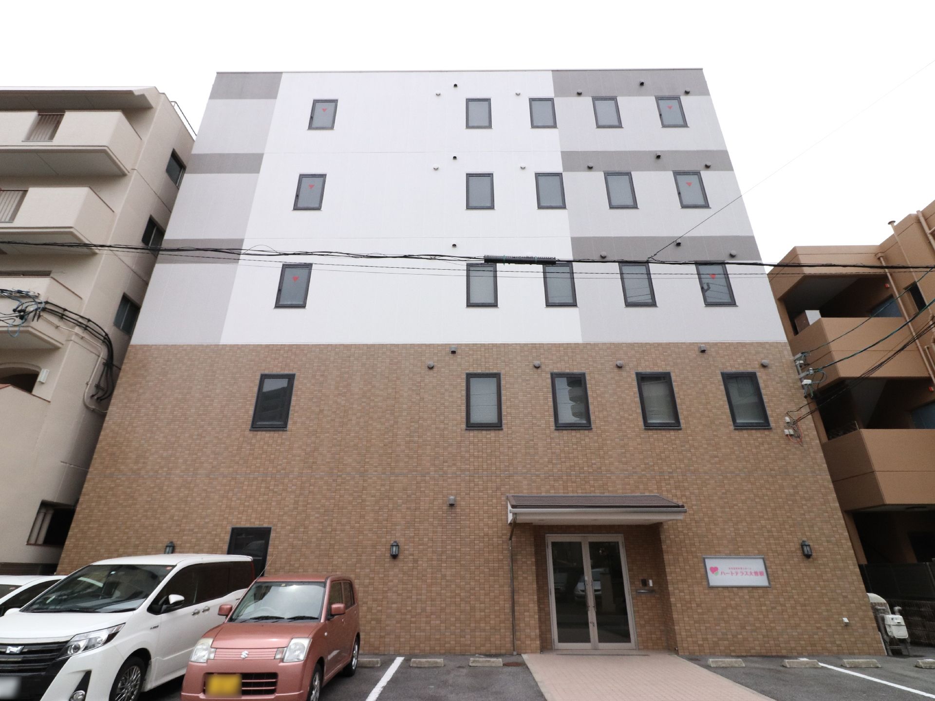 施設外観は茶色と白、グレーの配色になっている。階数によって色は違う。また施設入口近くは車の駐車スペースになっている。