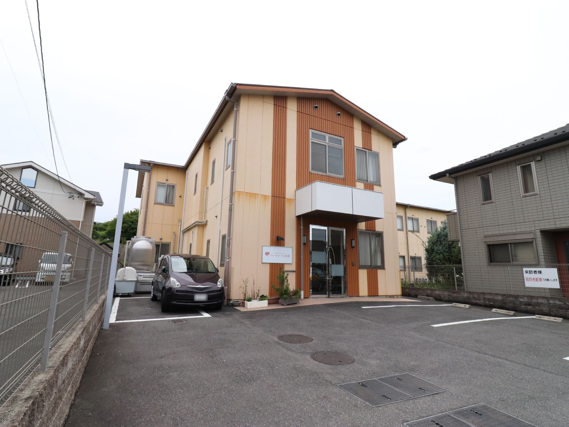 施設外観は茶系でまとめられている。入り口には屋根があり、施設の看板が取り付けられている。車の駐車スペースもある。
