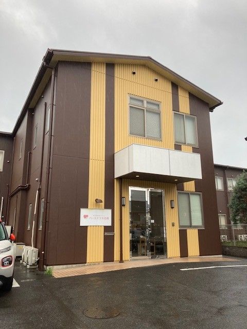 施設外観は茶系でまとめられている。入り口には屋根があり、施設の看板が取り付けられている。車の駐車スペースもある。