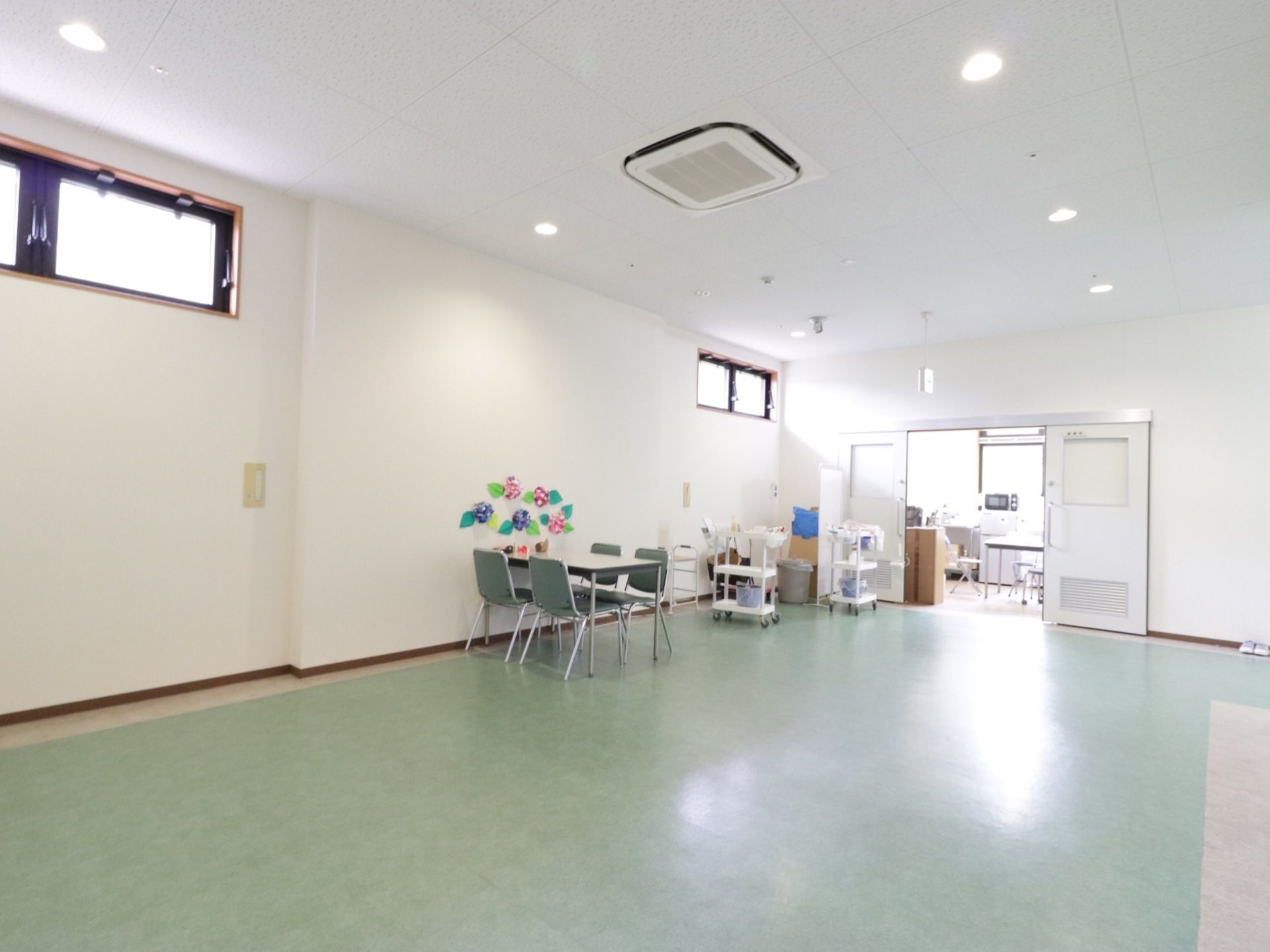 薄緑の床の広々とした空間。その壁際に４脚の椅子とテーブルのセットが設置されており、側には職員が使用するトレーが数台置かれている。