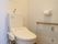 トイレには洋式便器が設置されている。壁にはセットされたトイレットペーパーと横長の手すり、呼び出しボタンがある。