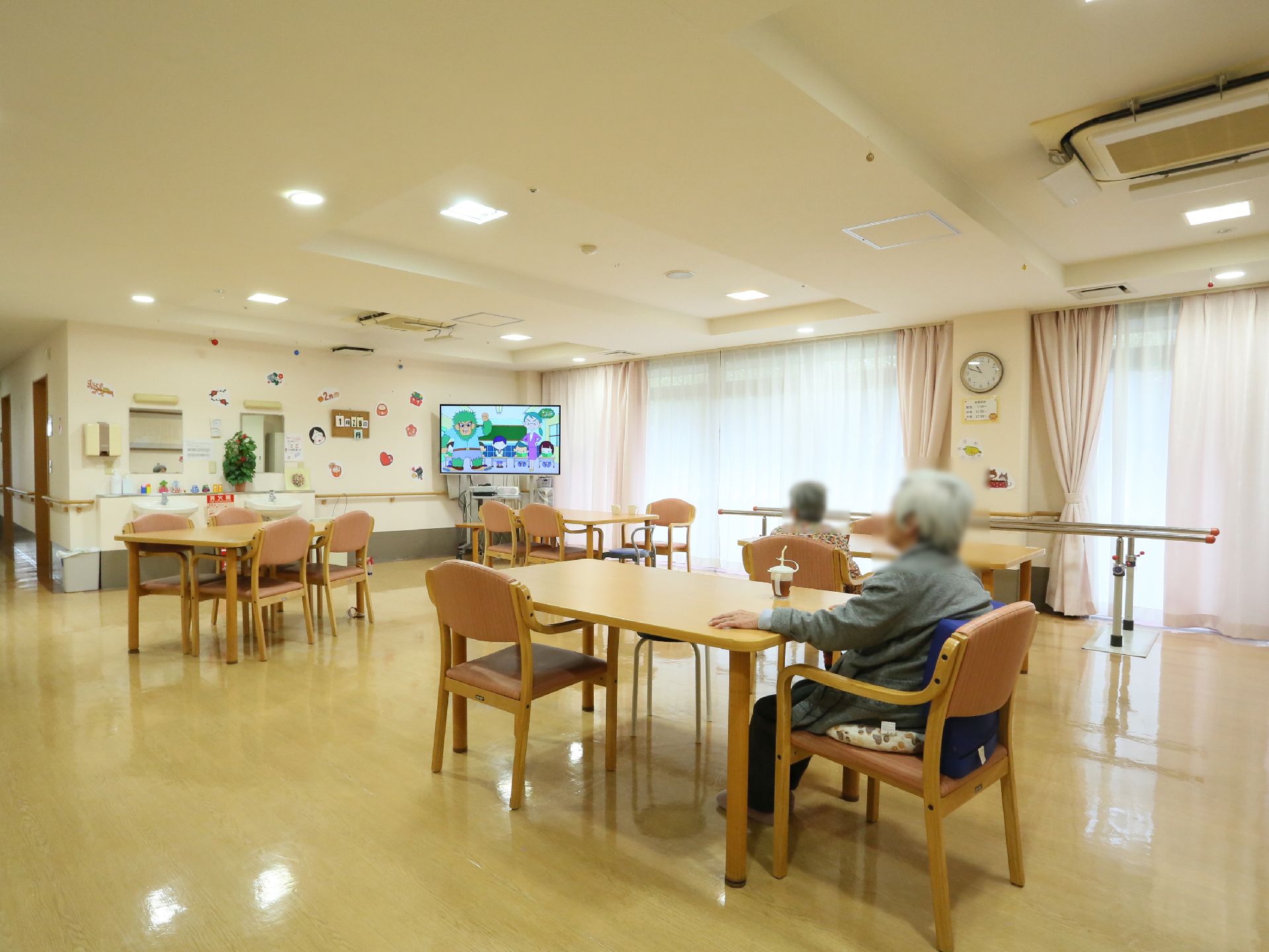 食堂にはテーブルと椅子が用意されている。椅子には入居者様が座ってテレビをご覧になっている。平行棒がある。