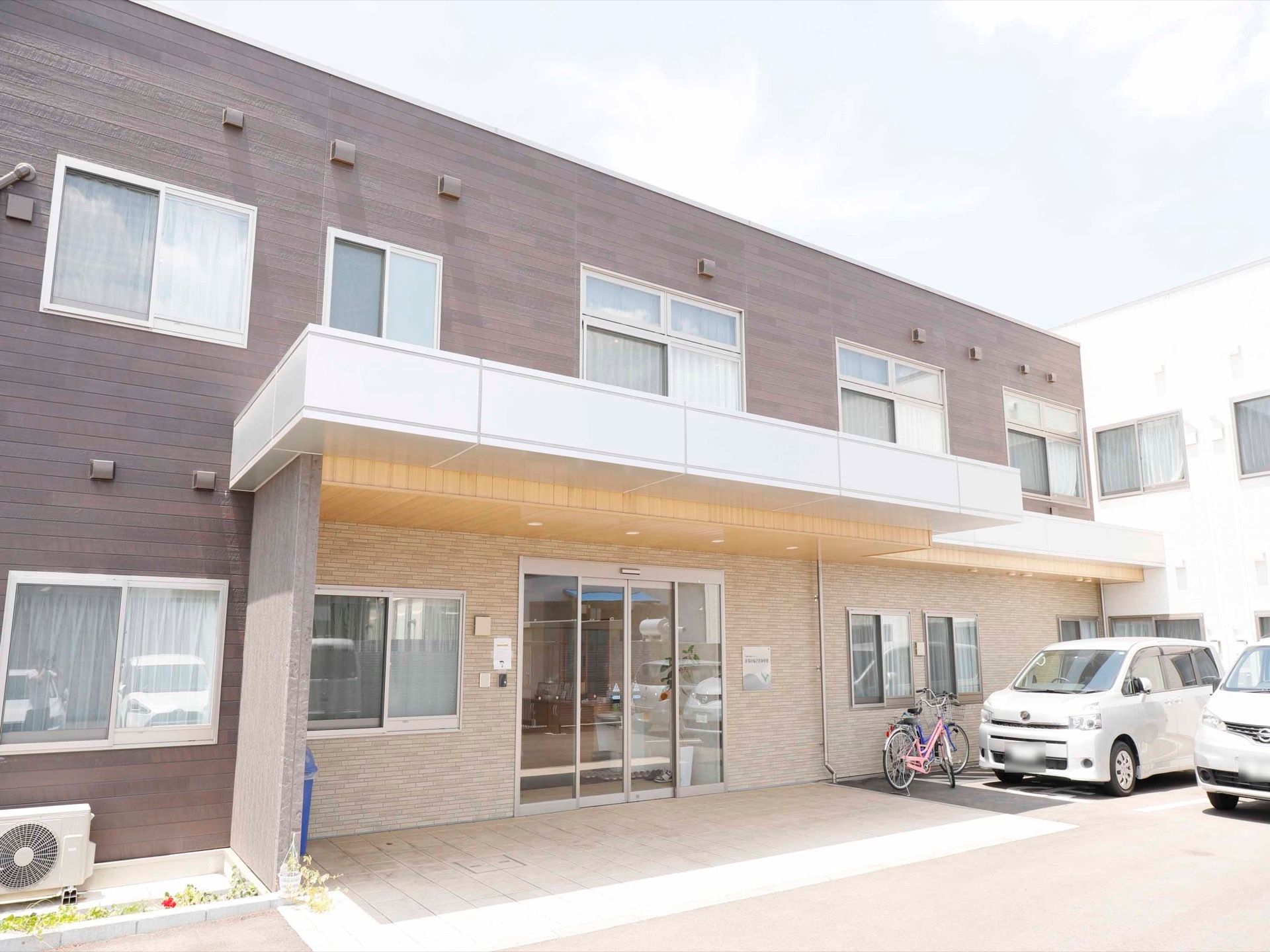 玄関は自動扉で、玄関ポーチは屋根がせり出している。入口横に自転車が置かれており、駐車場が並んでいる。