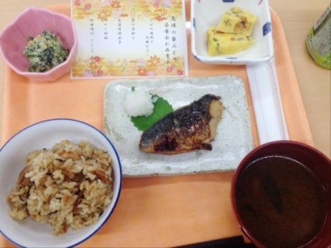 オレンジ色のトレーの上に炊き込みごはんと三つのおかず、お吸い物が置かれている。メニュー表も用意されている。