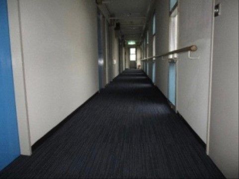 カーペット張りの廊下である。片側に青い扉の居室が並び、反対側には窓と手すりが設置されている。窓から日の光が差し込んでいる。