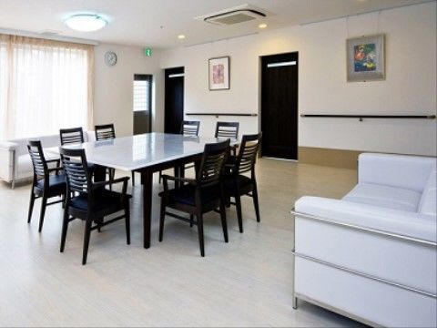 床と壁が白い部屋の真ん中に、大きめのテーブルとおしゃれな椅子が8脚置いてある。白いソファーもそばに置いてある。