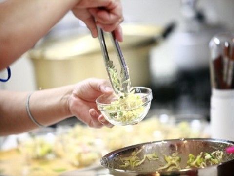 施設の厨房で調理している場面である。ボールの中でかき混ぜられた野菜をトングでつかみ、ガラス製の食器に移している。