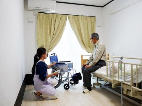 居室の大きな窓には、レモン色のセンタークロスのカーテンを掛けお洒落なイメージである。壁際に介護用ベッドを備え、天井にエアコンを設置している。