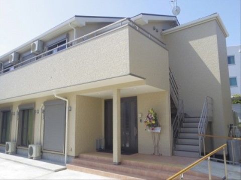 オフホワイトの２階建て建物の横に階段があり、お洒落な雰囲気である。玄関ドア横にお花を飾り、華やかな空間を設けている。